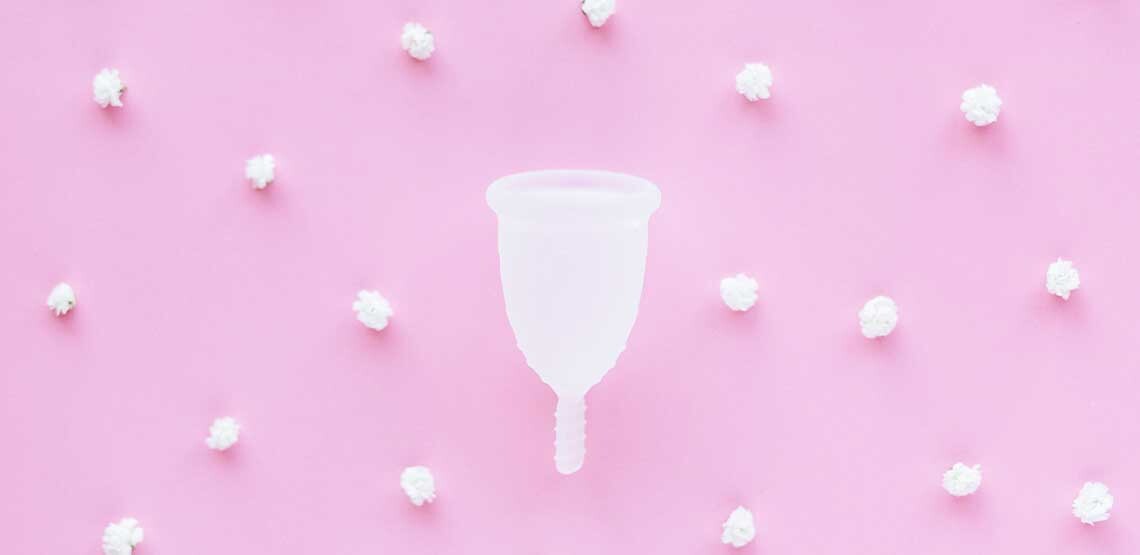 A picture of a white silicone period cup on a pink background and tiny, white cotton balls spread out in a pattern around the period cup.