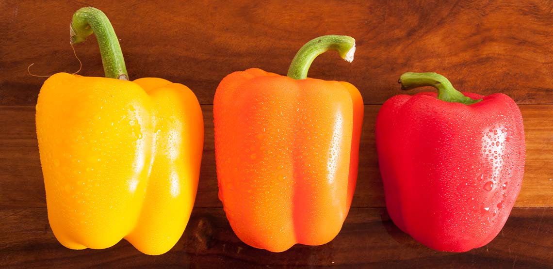 A yellow, orange and red pepper in a line on a wooden table.