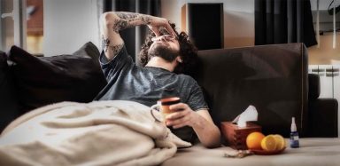 A man laying on a couch with a mug in his hand and holding his head.
