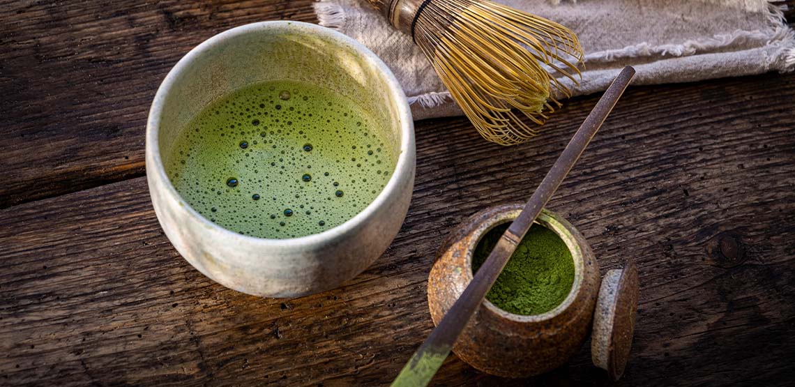 A small cup of matcha, a whisk and a container of matcha powder on a wooden table.
