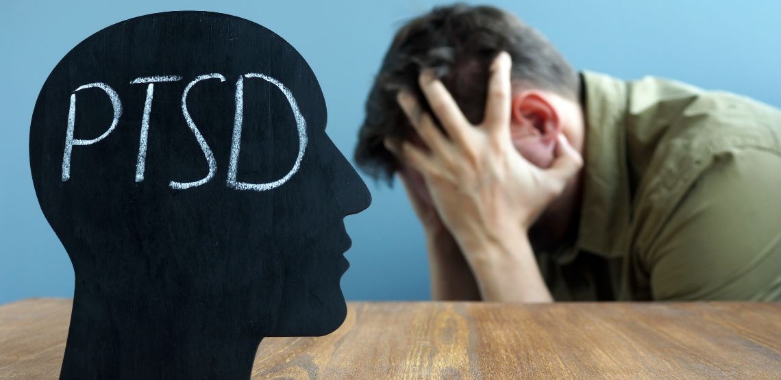 A cutout of a head with "PTSD" written on it, with a man in the background holding his head.
