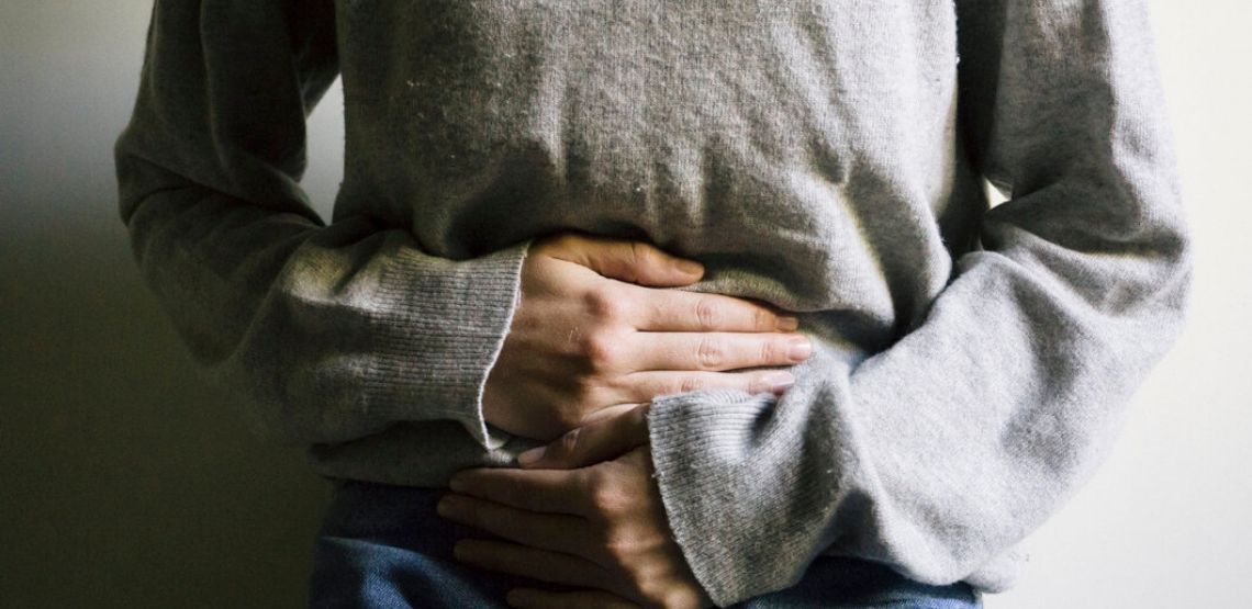 A close up of a woman in a grey sweater. The image is focused on her torso, with both hands holding her stomach.