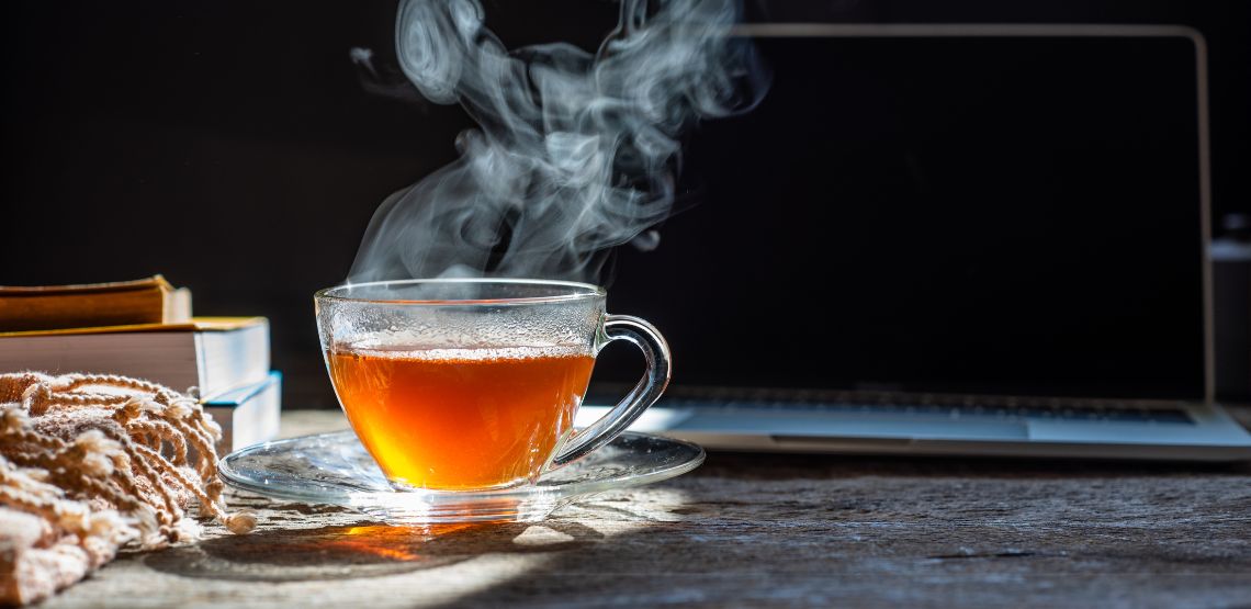 A steaming mug of black tea in front of a laptop. The mug is made of glass and is see-through.