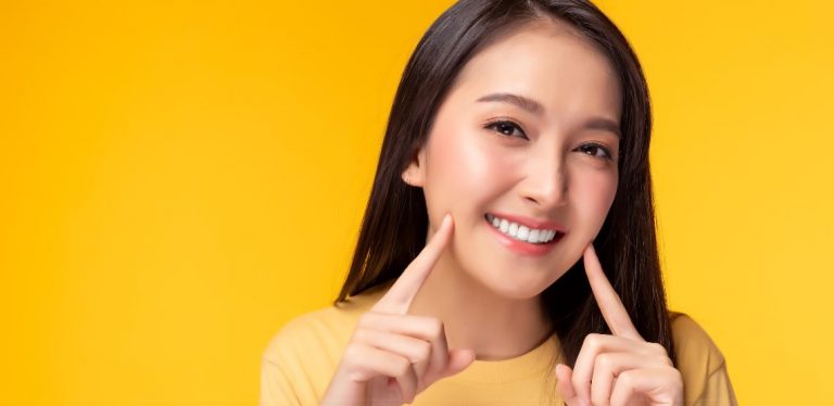 A woman smiling, pointing to her face. She is in front of a bright yellow background.